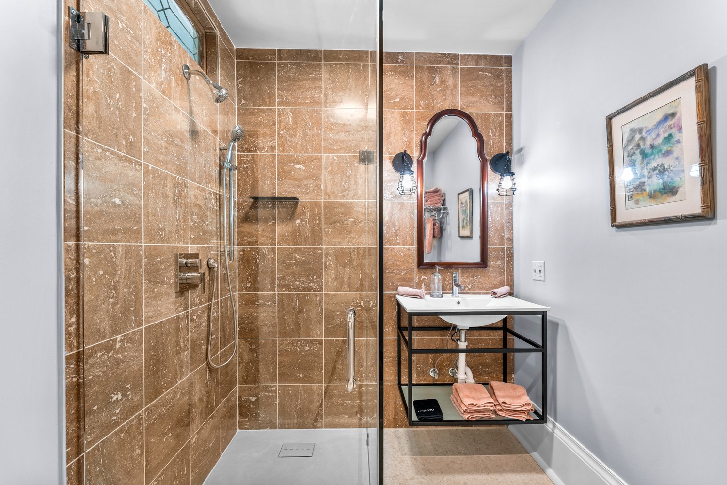 A bright bathroom featuring a large glass-enclosed shower with light brown tiled walls next to a modern sink and mirror flanked by wall sconces.