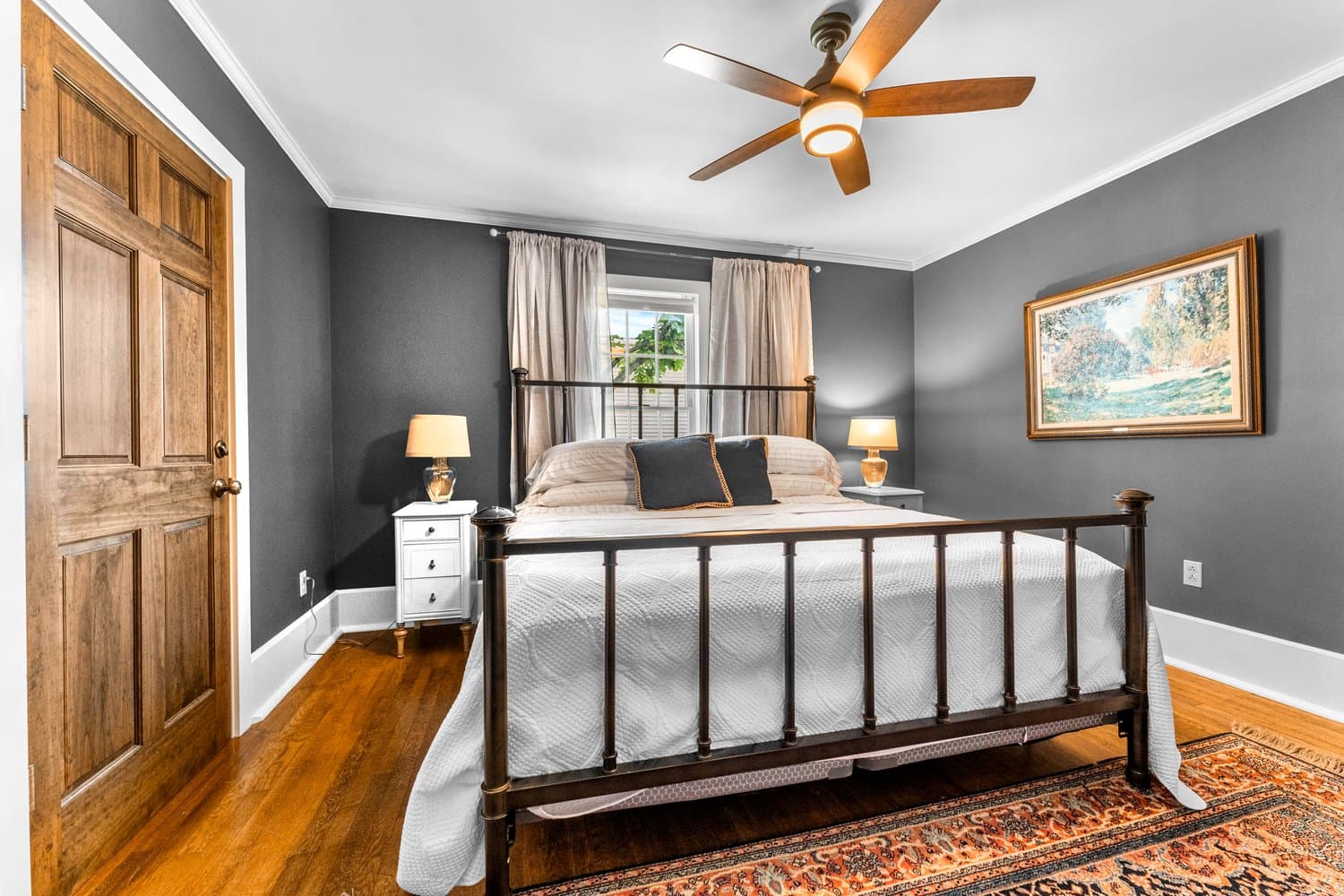 Pristine bedroom with metal-framed king-size bed flanked by nightstands with table lamps, hardwood floors, and ceiling fan.
