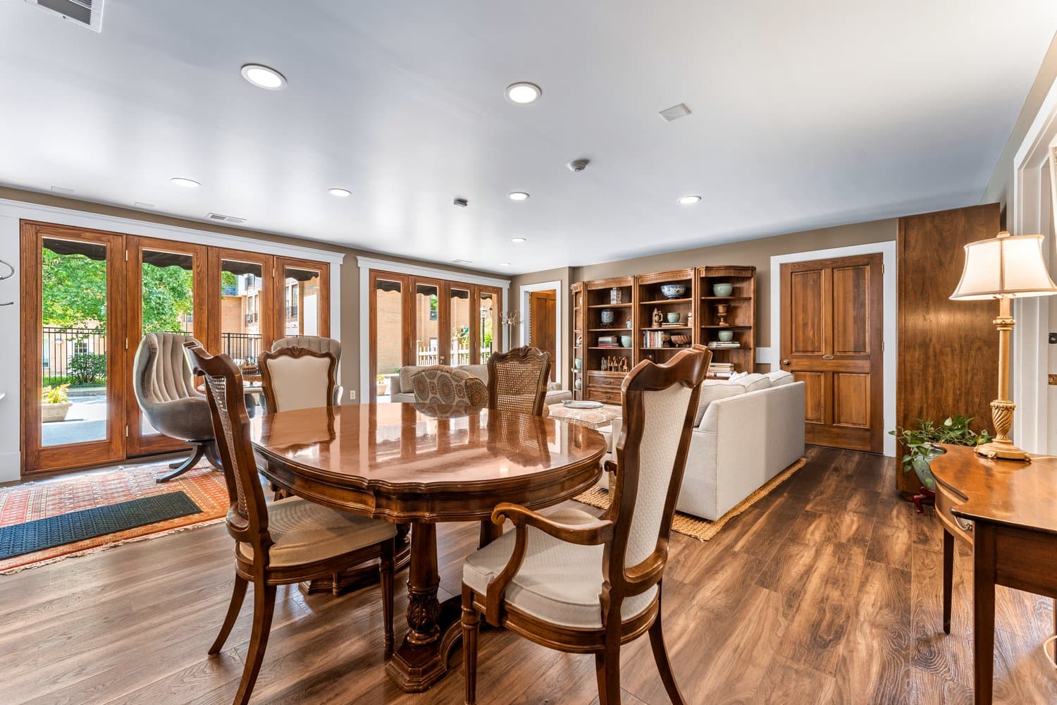 Elegant dining area with traditional table and chairs for four next to a wall of sunny windows, and living area visible in the background.