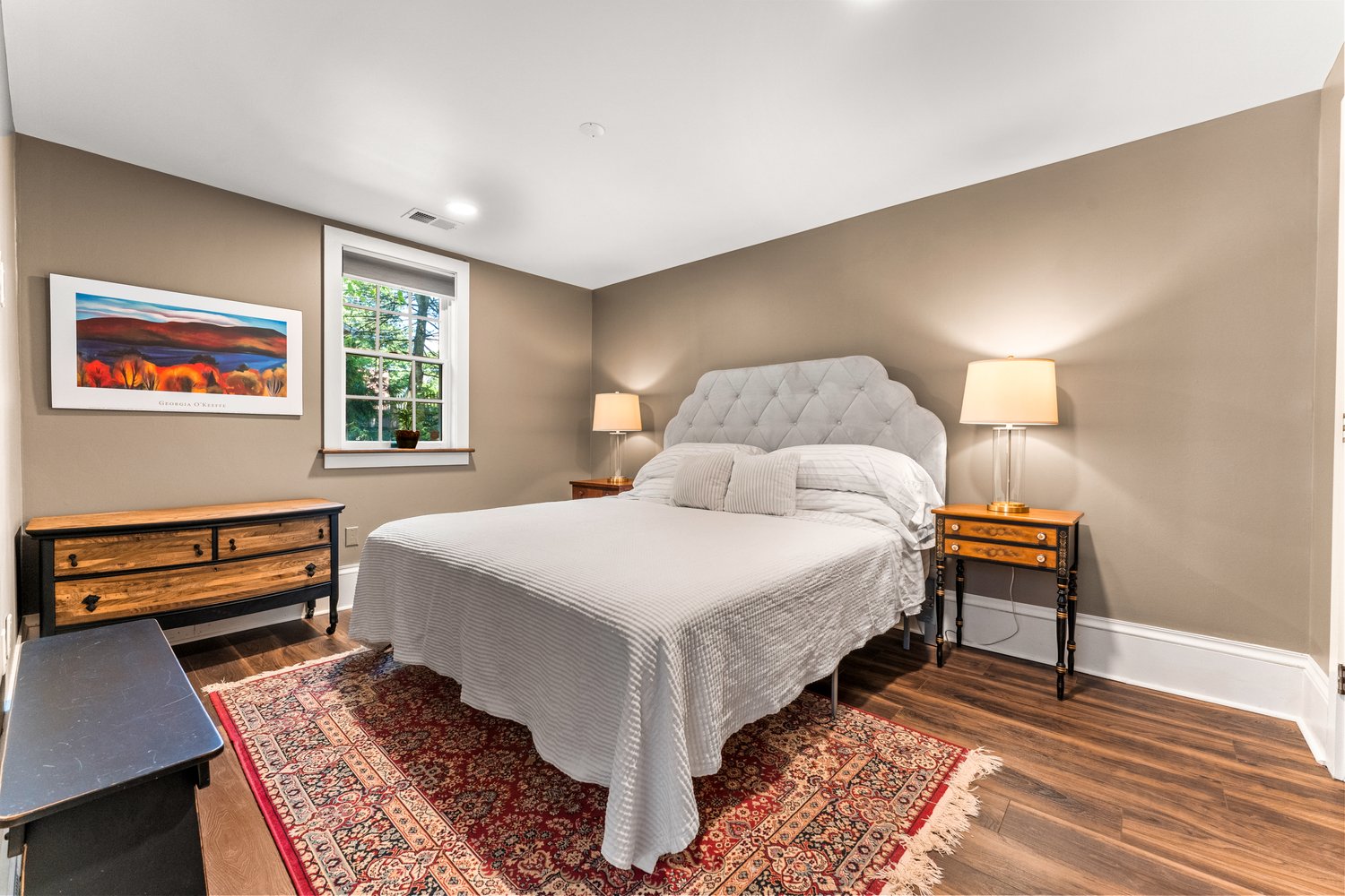Inviting bedroom with king bed with upholstered headboard and all white linens, hardwood floors, and oriental area rug.