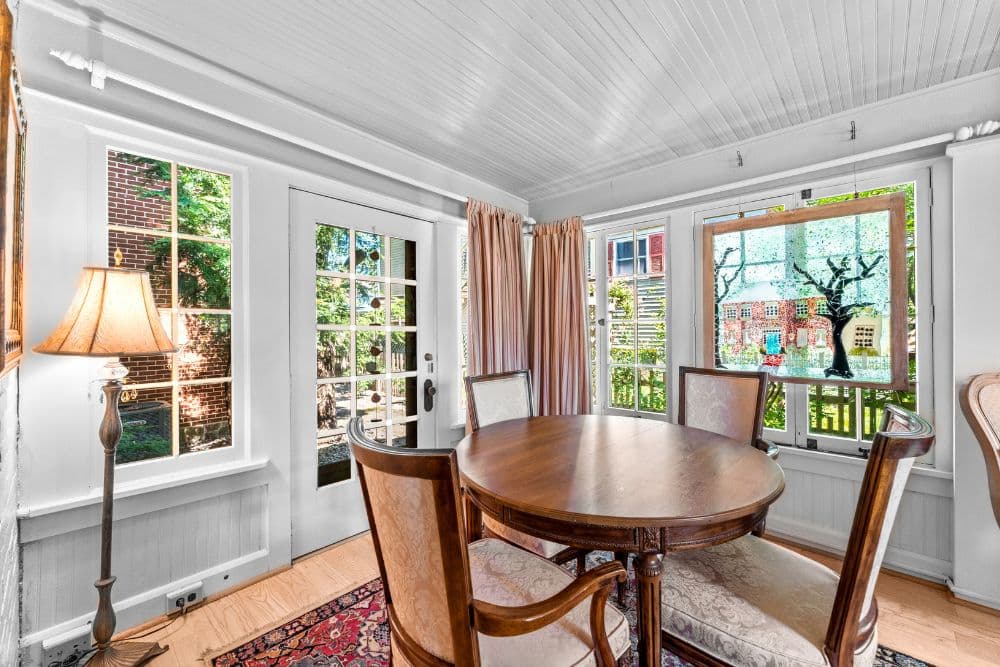 Bright and airy dining area featuring a round wooden table, elegant chairs, and large windows with vibrant greenery outside.