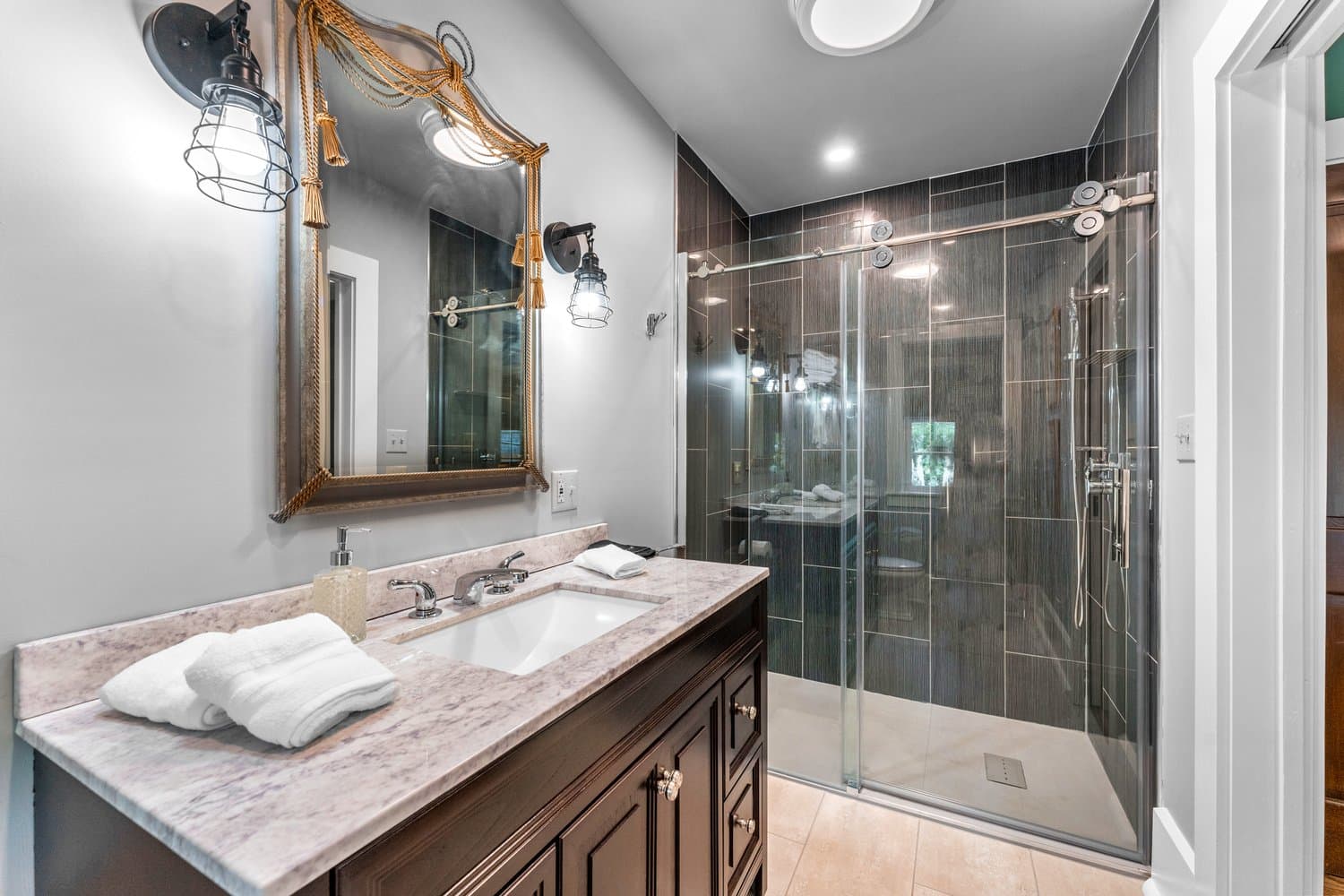 An elegant modern bathroom with large glass-enclosed tiled shower, sink with vanity, and mirror flanked by sconces.