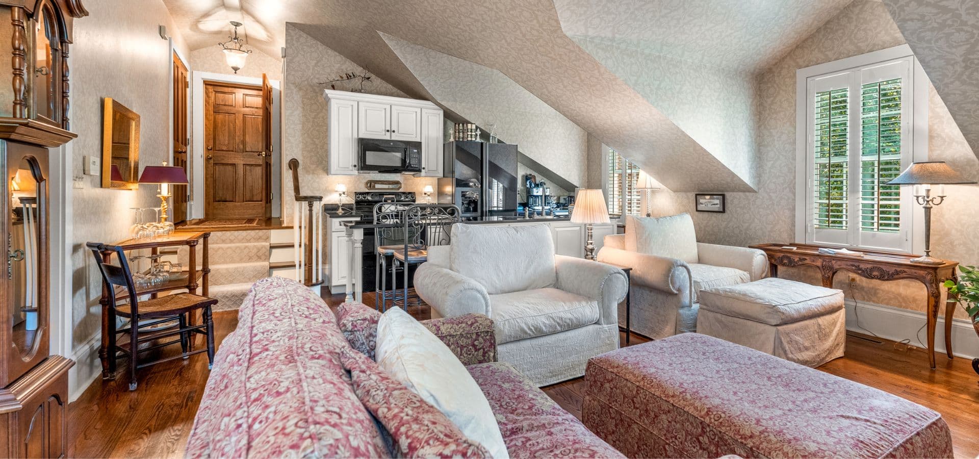 A cozy living room features white sofas, a patterned armchair, and a small kitchen area under sloped ceilings.