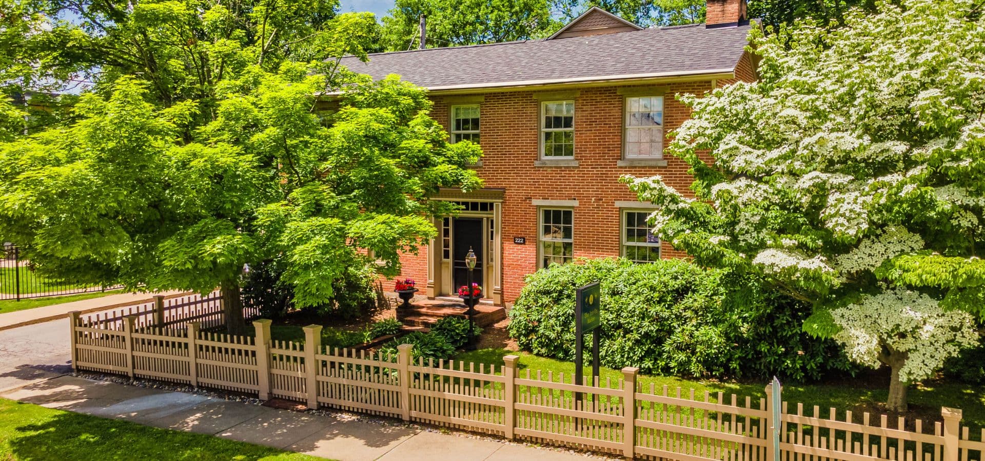 A brick house surrounded by lush greenery and a white picket fence on a sunny day.