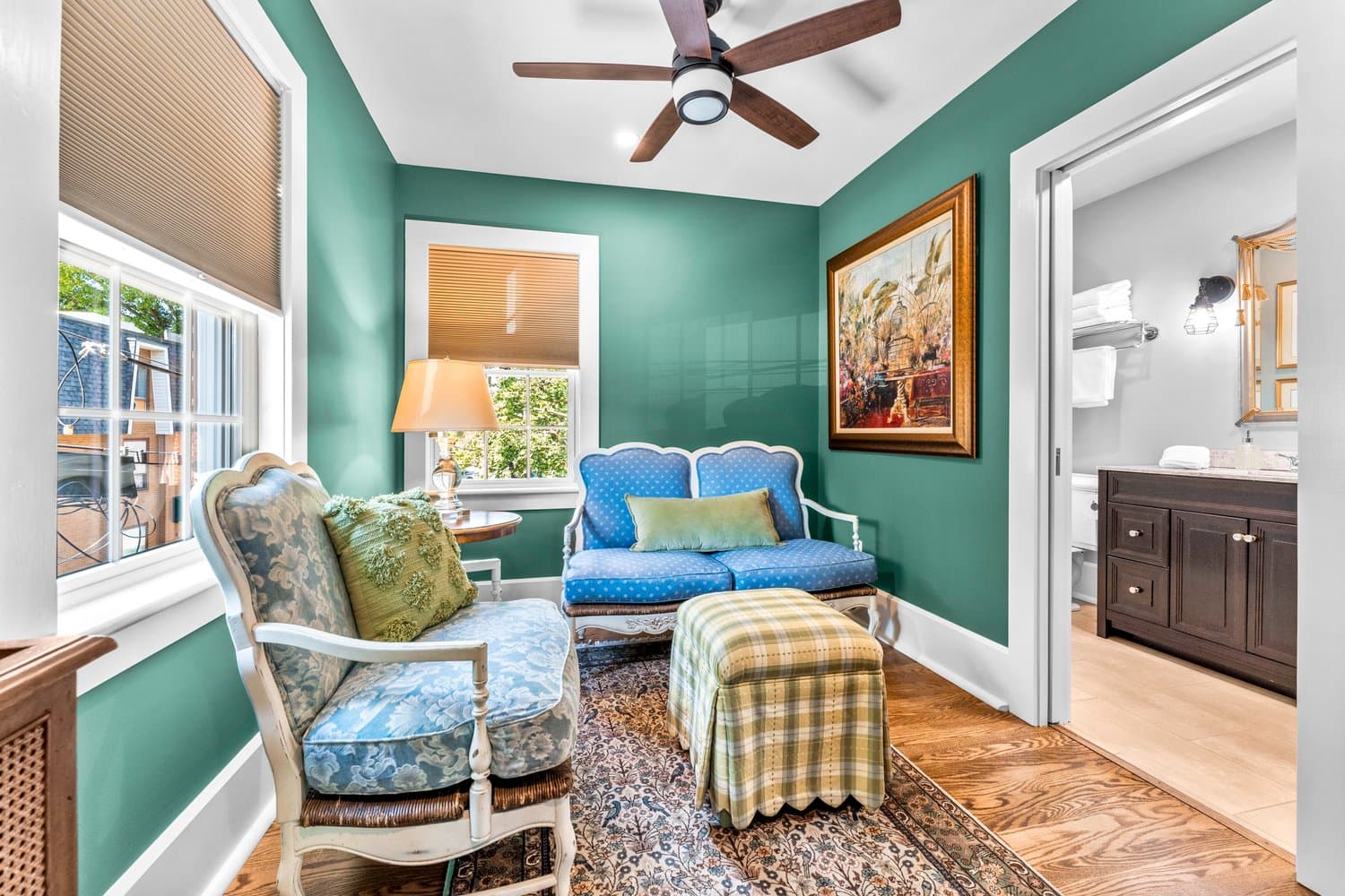 A charming sitting area with lots of natural light, two wooden white painted loveseats with thick floral cushions, a skirted ottoman, a ceiling fan, and hardwood floors.