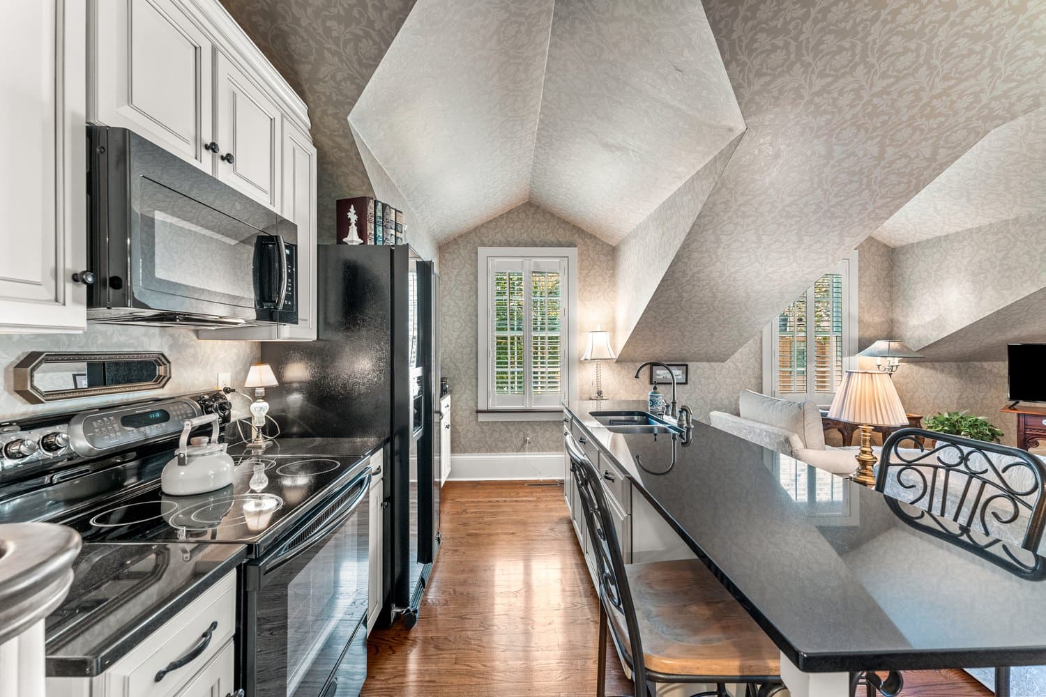 A well-lit kitchen under a slanted ceiling with white walls and cabinetry, stainless appliances, and a kitchen island with prep sink.