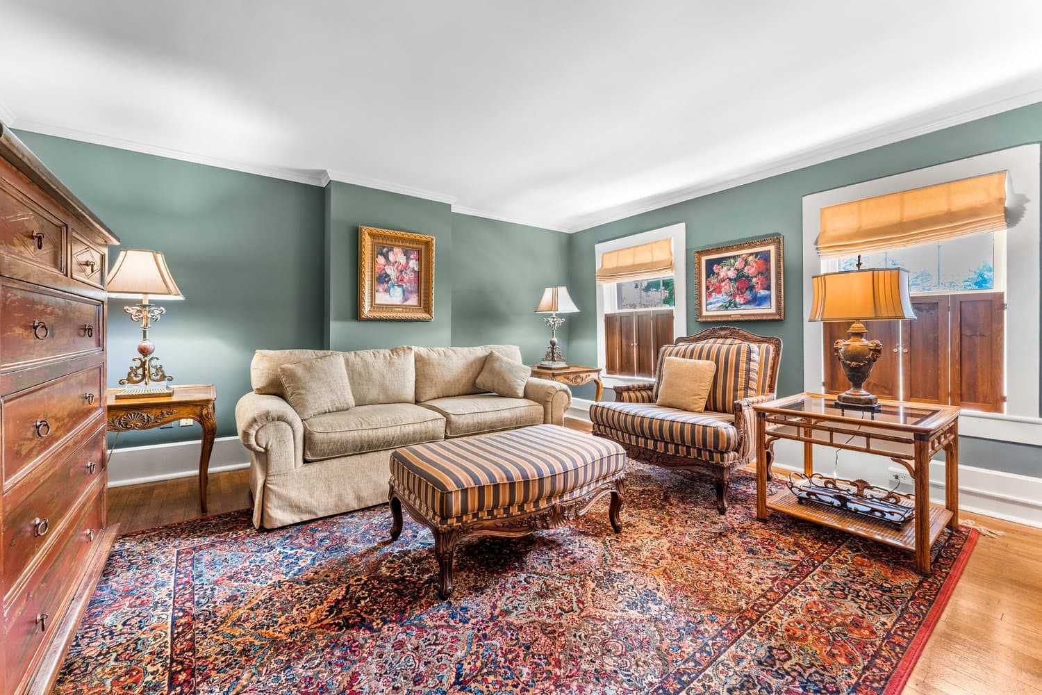 Large, well-lit, and inviting living room with traditional furnishings including deep-cushioned sofa, armchair, and ottoman, hardwood floors, and oriental area rug.