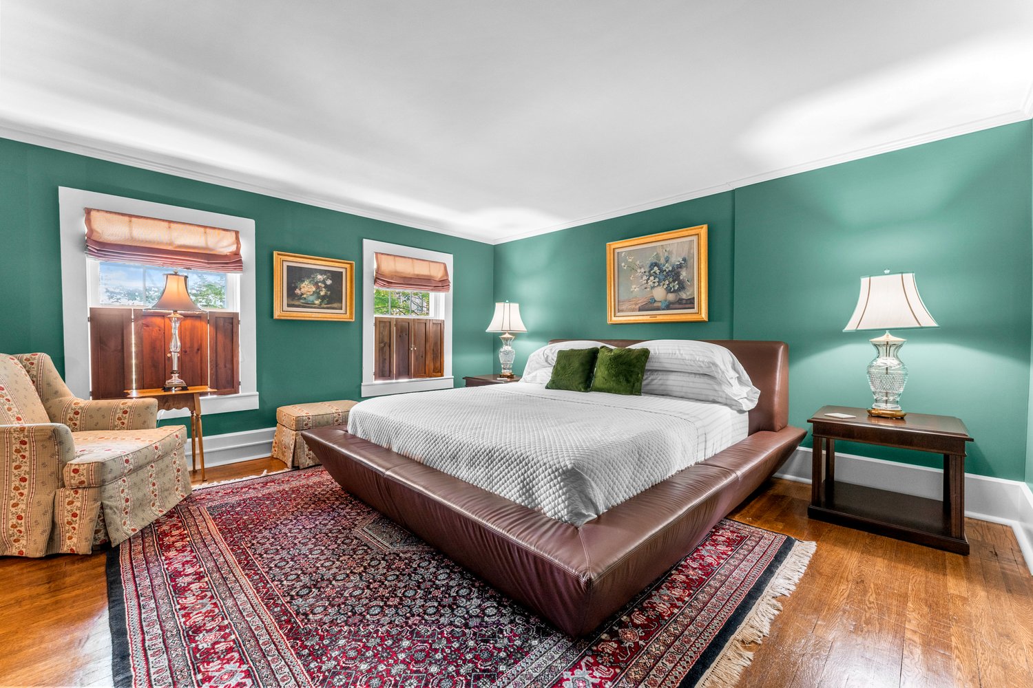 A brightly lit bedroom with teal walls, hardwood floors, and a modern platform bed with a dark leather frame dressed in white linens and green pillows.