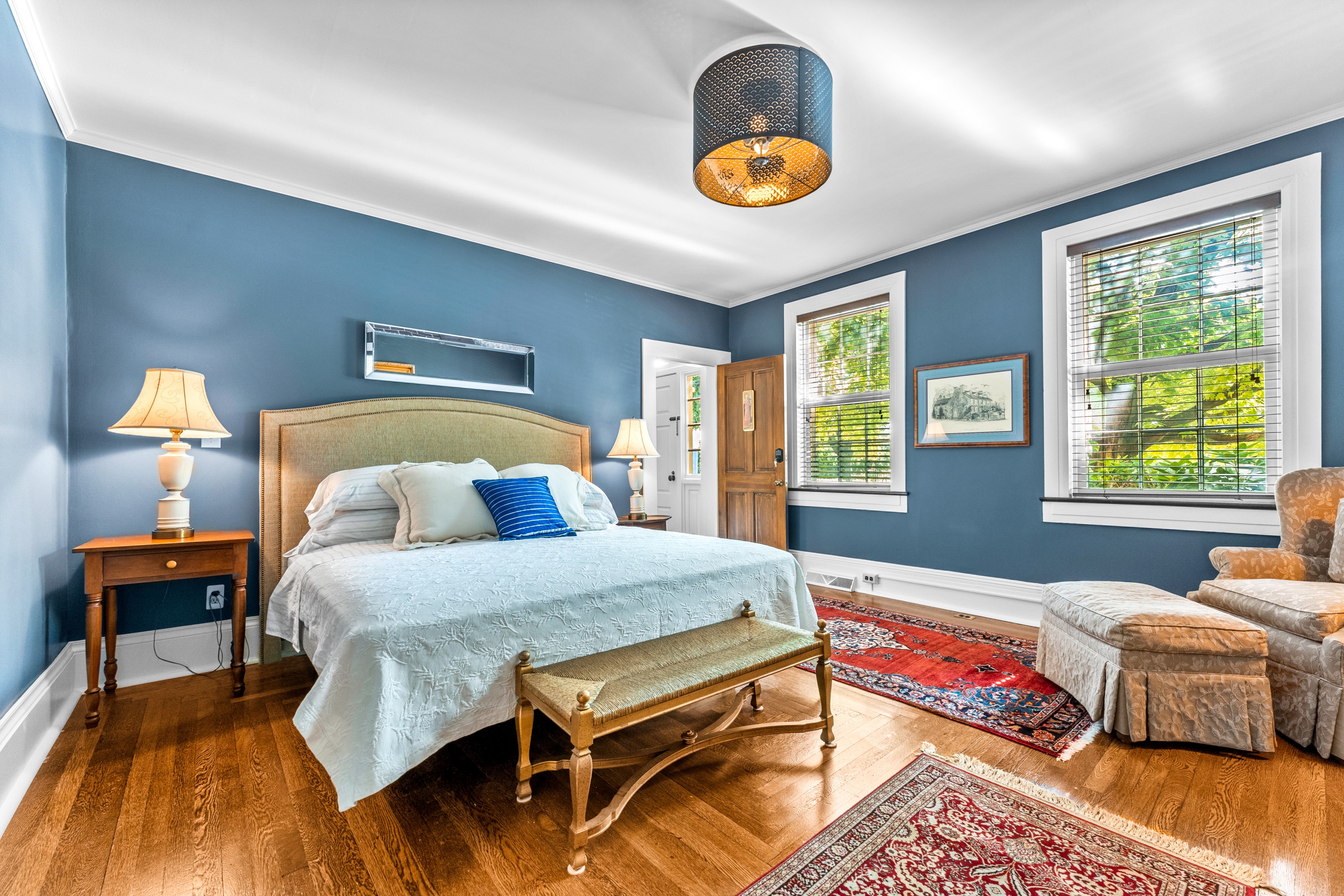 A well-lit bedroom with blue walls, hardwood floors, and a king bed dressed in white linens with a blue accent pillow.