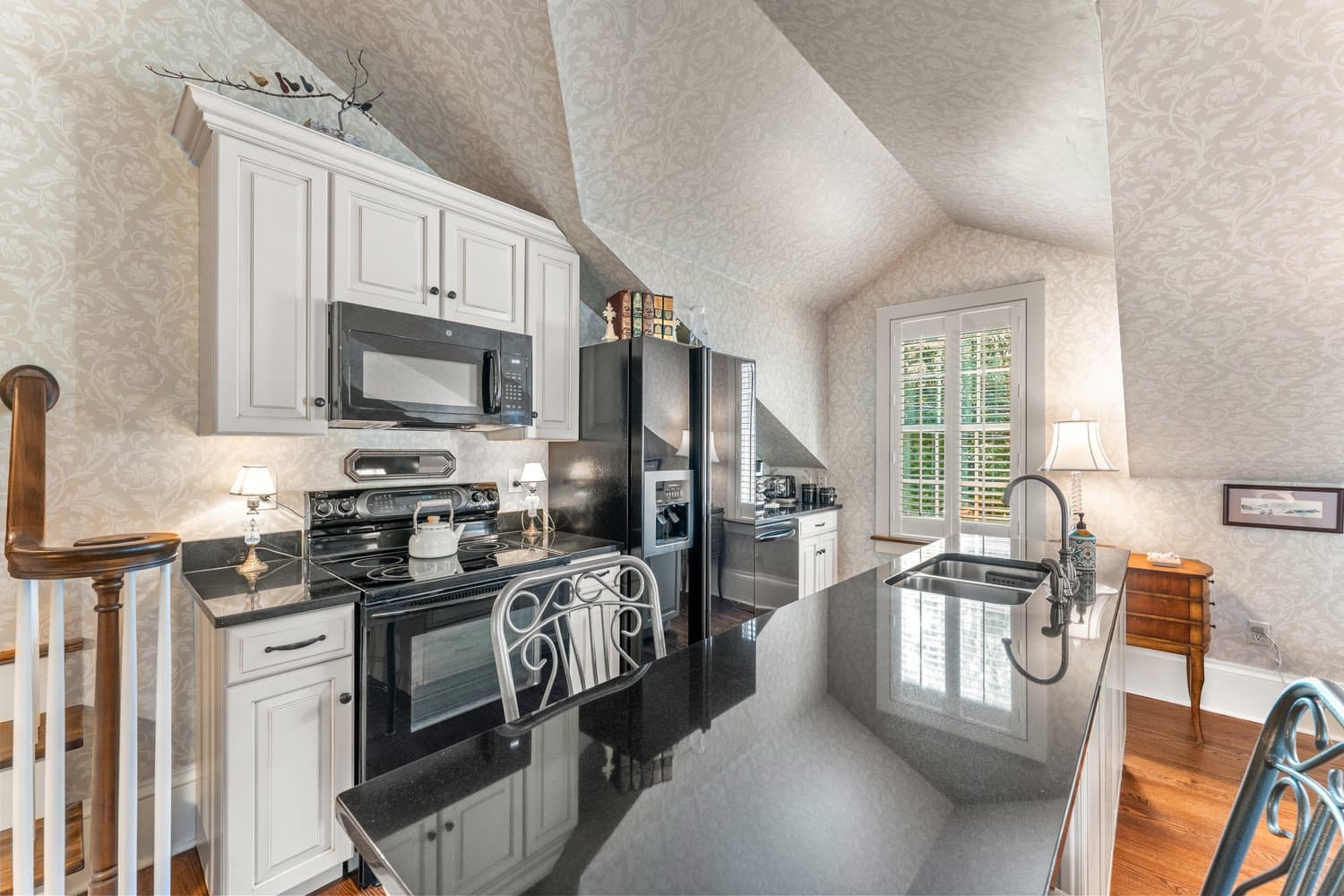 A well-lit kitchen under a slanted ceiling with white walls and cabinetry, stainless appliances, and a kitchen island with prep sink.