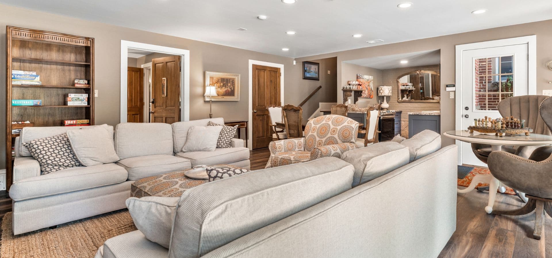 A cozy living room featuring a light gray sectional sofa, patterned accent chairs, a wooden bookshelf, and warm lighting.